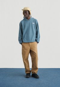 Μπλε φλις φούτερ με κουκούλα και λογότυπο μπροστά, χαλαρή παντελόνι σε καφέ απόχρωση και μαύρα αθλητικά παπούτσια. Φοράει κίτρινο καπέλο τύπου κουβά με καλύμματα για τα αυτιά.