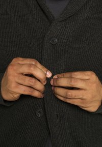 Dunkel gestrickte Strickjacke mit einem Schalkragen, die aus strukturiertem Stoff besteht und zwei schwarze Knöpfe hat. Hände schließen den vorderen Verschluss.