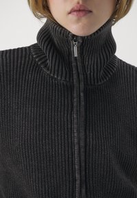 Schwarzer, gerippter Strickzipper mit hohem Kragen und kontrastierender Textur am Hals. Mit einem silbernen Reißverschluss.