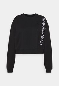 Sudadera negra recortada con mangas largas, que presenta "CALVIN KLEIN JEANS" en texto blanco en negrita en la manga izquierda y un cuello redondo clásico.