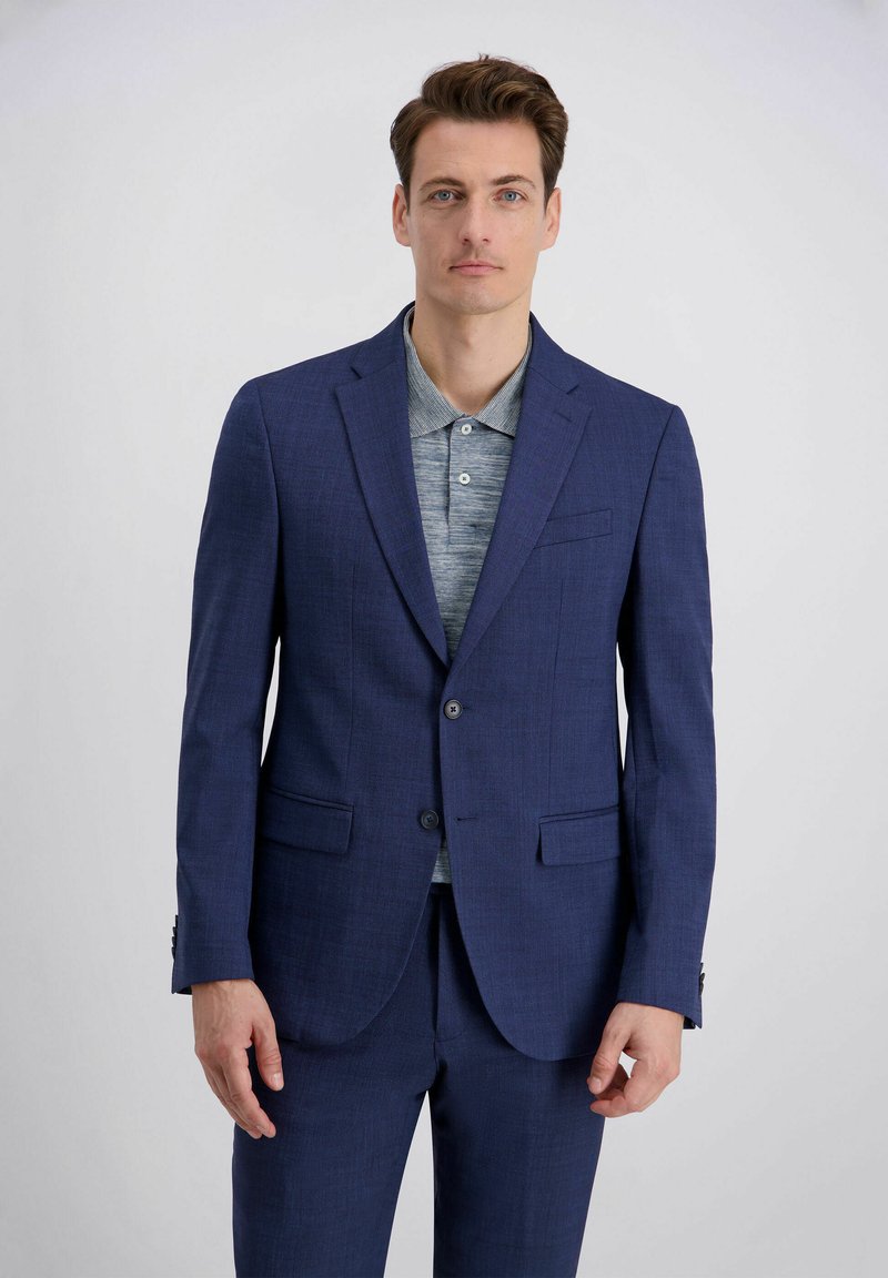 Mann in einem maßgeschneiderten marineblauen Anzug und grauem Poloshirt vor einem schlichten hellen Hintergrund stehend.