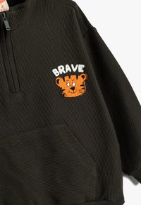 Suéter negro con canalé, con cremallera y bolsillo frontal, que presenta una cara de tigre naranja bordada y la palabra "BRAVO" en blanco encima.