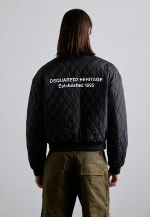 Veste bomber matelassée noire avec des poignets côtelés, ornée de texte blanc au dos : "DSQUARED2 HERITAGE Établi en 1995."