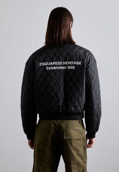 Veste bomber matelassée noire avec des poignets côtelés, ornée de texte blanc au dos : "DSQUARED2 HERITAGE Établi en 1995."