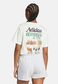 Light-colored cropped t-shirt with "Adidas" logo and graphic of a market on the back; paired with gray shorts. Cotton material, casual design.