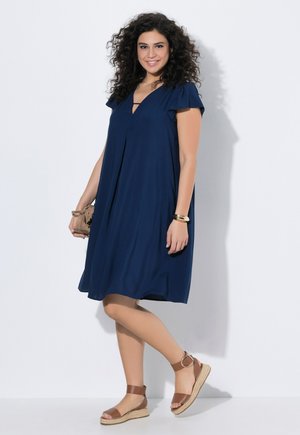 Frau mit lockigem Haar, die ein knielanges, marineblaues Kleid und braune Sandalen mit Knöchelriemen trägt, hält eine beigefarbene Clutch und lächelt.