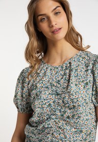 Blusa floreale con maniche a sbuffo corte, realizzata in tessuto azzurro chiaro con motivi di margherite bianche e fogliame verde. Dettaglio arricciato nella parte anteriore.