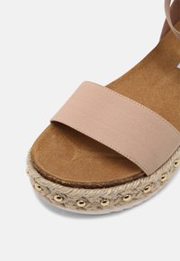 Gros plan d'une sandale beige à bride avec une semelle intérieure en liège, une semelle plateforme en corde tressée et de petits clous dorés autour du bord de la semelle.