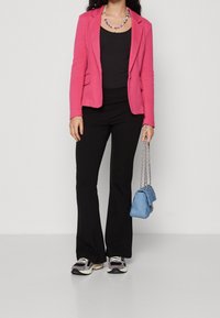 Rosa blazer, svart topp, svarta utsvängda byxor och grå sneakers. Accessoar: liten blå handväska med kedja. Släta tygtexturer.