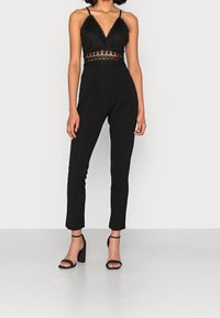 Kvinna i en ärmlös svart jumpsuit med spetsdetalj i midjan och svarta högklackade sandaletter med ankelsnöre, stående mot en neutral bakgrund.