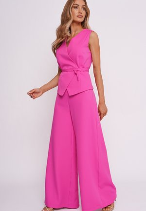 Kobieta z długimi falującymi włosami, ubrana w bezrękawnik w jasnoróżowym kolorze i spodnie o szerokich nogawkach, stoi na tle jednolitego jasnego tła.