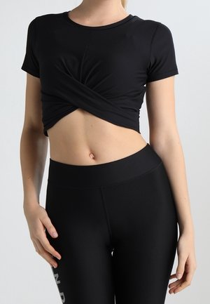Femme portant un crop top noir torsadé et des leggings noirs taille haute, debout devant un fond clair uni.