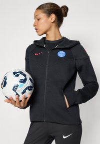 Svart zip-up jakke med hette og sidelommer. Har en blå Paris-logo og rød Nike swoosh. Modellen holder en hvit fotball med blå detaljer.