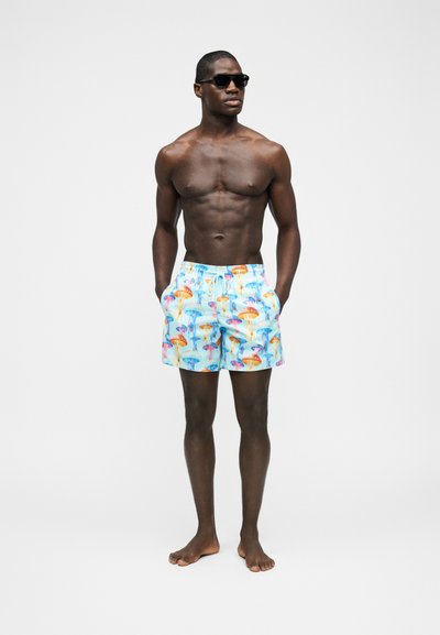 Μυώδης άντρας φοράει σκούρα γυαλιά ηλίου και μπλε μαγιό με πολύχρωμη εκτύπωση μέδουσας, στέκεται ξυπόλυτος με τα χέρια στις τσέπες.