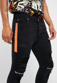 Jeans ajustados negros con detalles desgastados, franja lateral naranja y bolsillo con cremallera; presenta una textura acanalada y acentos deshilachados.