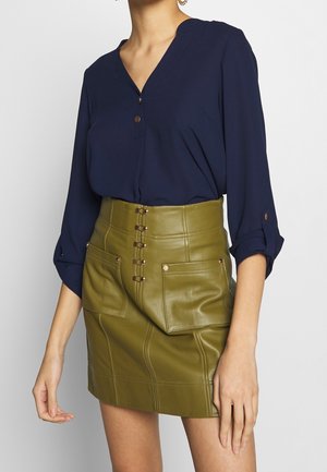 Femme portant un chemisier bleu marine à col en V avec boutons et manches retroussées, associé à une jupe en cuir vert olive taille haute avec poches avant et fermoirs.