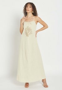 Mujer con cabello rizado, vestida con un vestido largo beige con bordado coral, tacones de tiras transparentes y pendientes dorados, posando con una mano en la cadera.