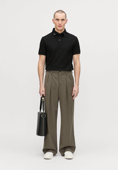 Black polo shirt with collar accents, high-waisted grey trousers, and white sneakers. Holding a cylindrical black bag with a strap.