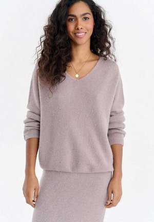 Młoda kobieta o długich, kręconych włosach, uśmiechająca się, ubrana w miękki, wrzosowy sweter i dopasowaną spódnicę, na jednolitym białym tle.