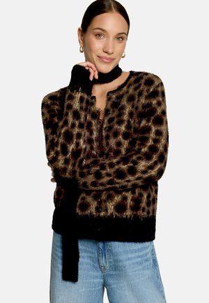 Femme aux cheveux foncés portant un cardigan duveteux à imprimé léopard, une écharpe noire, des boucles d’oreilles anneaux dorées et un jean bleu clair, souriante avec la main près du visage.