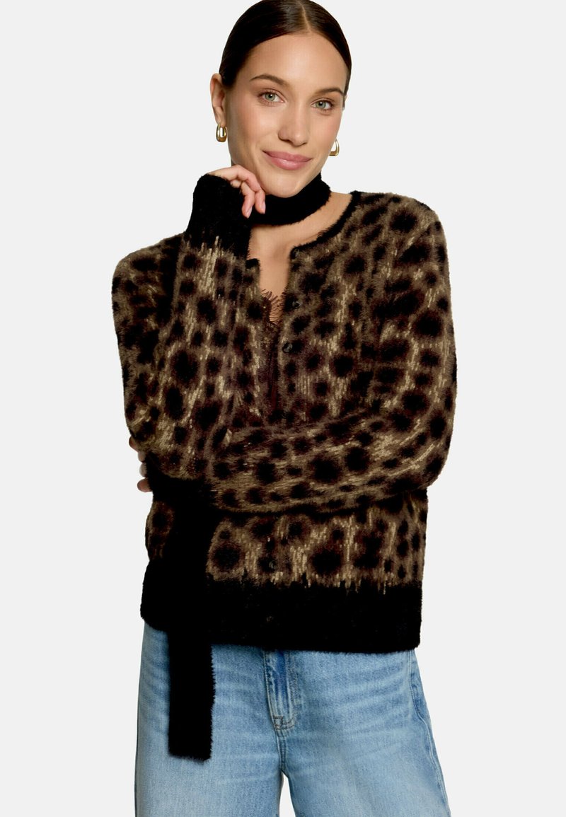 Femme aux cheveux foncés portant un cardigan duveteux à imprimé léopard, une écharpe noire, des boucles d’oreilles anneaux dorées et un jean bleu clair, souriante avec la main près du visage.
