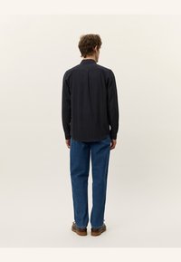 Schwarzes Langarmshirt mit strukturierter Stoffqualität, geradem Saum und Knopfdetails, kombiniert mit blauen Jeans im geraden Schnitt und braunen Schuhen.