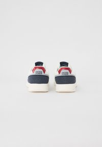 White sneakers with navy blue heel, red and gray accents. Smooth upper with textured heel, rubber sole. Branding on the back.