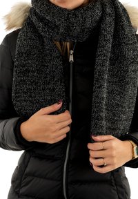 Scarf chunky nera e grigia lavorata a maglia, realizzata in morbido filato testurizzato, avvolta attorno al collo, in contrasto con un giubbotto imbottito nero.