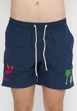 Uomo che indossa pantaloncini da bagno blu navy con coulisse bianca, fiore di ibisco rosso sul lato sinistro e ricamo a palma sul lato destro.