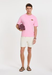 Camiseta de algodón rosa con un pequeño gráfico, pantalones cortos crema y sandalias marrones. Sosteniendo una toalla enrollada a rayas burdeos.