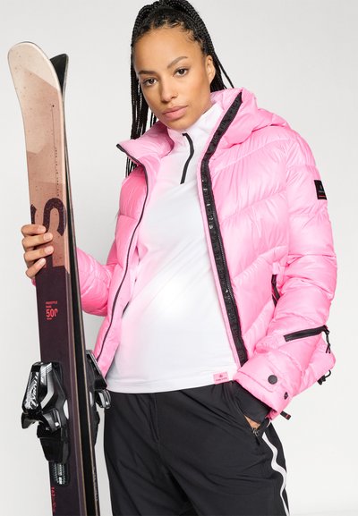 Γυναίκα σε ροζ μπουφάν holding μαύρα και ροζ σκι, φορώντας λευκή μπλούζα με φερμουάρ και μαύρο παντελόνι με λευκές ρίγες στην πλευρά.