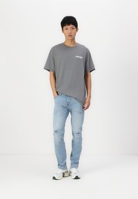 Szara bawełniana koszulka z logo "Wrangler", połączona z jasnoniebieskimi dżinsami z przetarciami i białymi trampkami z zielonymi akcentami.