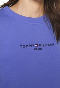 Fioletowy bawełniany sweatshirt z prążkowanym dekoltem i haftowanym logo marki. Tekst zawiera "TOMMY HILFIGER EST. 1985" oraz detal z blokiem kolorów.