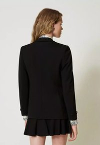 Zwarte blazer met gestructureerd ontwerp, met inkepingen bij de revers en knoopmanchetten. Gelaagd over een geblokt shirt, gecombineerd met een uitlopende rok.