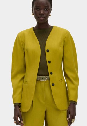 Donkere huidkleurige persoon die een gele blazer en broek draagt met zwarte knopen, een olijfgroene geribbelde top, bijpassende riem en gouden hoepeloorbellen.