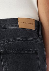 Black denim jeans featuring a tan leather tag with "SAM Søe" stamped on it, visible stitching, and a fitted design around the waist.