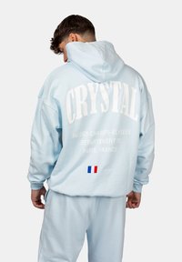 Hellblaues Hoodie mit weißem "CRYSTAL"-Text und Paris-Standortdruck auf der Rückseite. Verfügt über eine Kängurutasche und gerippte Bündchen.