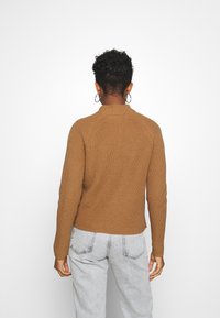 Personne aux cheveux courts et bouclés portant des boucles d'oreilles créoles, un pull marron à côtes et un jean gris clair, vue de dos sur un fond blanc.