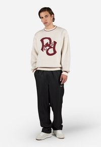 Hvid striktrøje med maroon logo-design, parret med løse sorte bukser. Trøjen har ribbede mansjetter og halsudskæring. Hvide sneakers.