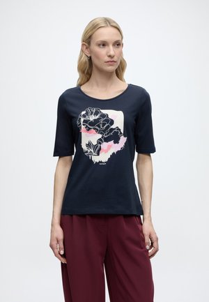 Kobieta z długimi blond włosami, ubrana w granatowy t-shirt z kwiatowym wzorem i bordowe spodnie, stojąca na tle jednolitego białego tła.