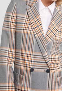 Dubbelgeknoopte blazer in een gemêleerde stof met grijs, zwart en oranje vierkanten. Opvallende details zijn onder andere twee zwarte knopen en een notch revers.
