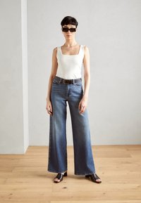 Modèle portant un débardeur blanc à encolure carrée, un jean en denim bleu à jambes larges, une ceinture noire et des sandales à talons noirs, se tenant sur un sol en bois.