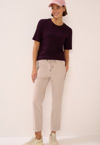 Chemise violette à manches courtes avec un col rond, associée à un pantalon beige clair à cordon de serrage et des baskets beiges, dotées de poches zippées sur les côtés.