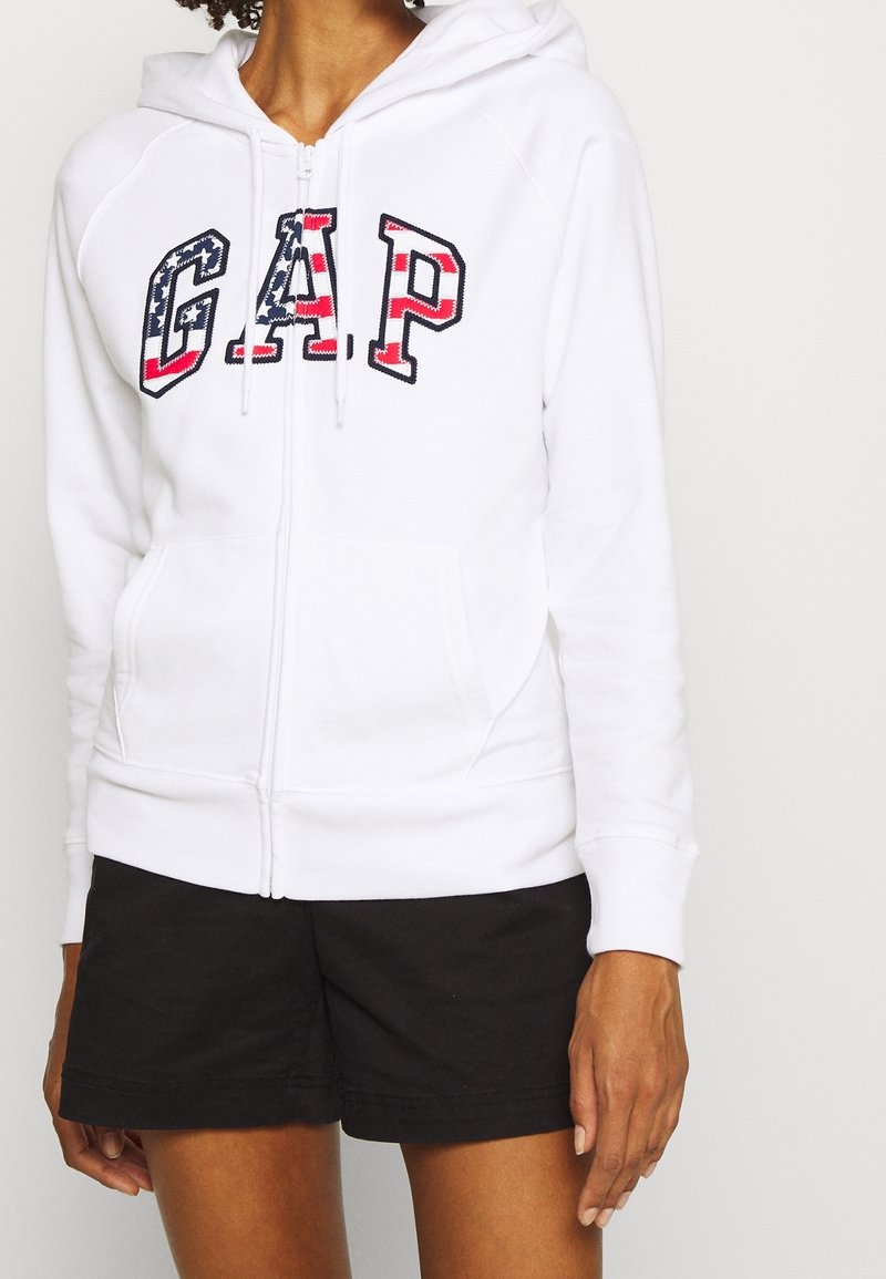 Persona vistiendo una sudadera blanca con cremallera que dice "GAP" en colores de la bandera americana y pantalones cortos negros, de pie contra un fondo simple.