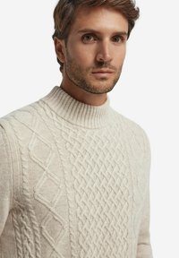 Sweter w kolorze kremowym, wykonany z dzianiny, z wysokim prążkowanym kołnierzem, z teksturowanymi wzorami w warkocze na całej powierzchni. Gładka faktura przypominająca wełnę.