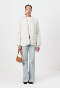 Chaqueta acolchada gris claro con rayas verticales, bolsillos frontales, llevada sobre una blusa color crema, combinada con jeans de campana azul claro y un bolso marrón.