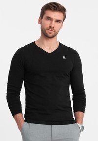 Schwarzes langärmliges V-Ausschnitt-Shirt mit einem figurbetonten Design, weichem Material und einem kleinen Logo auf der Brust, getragen über hellgrauen Hosen.