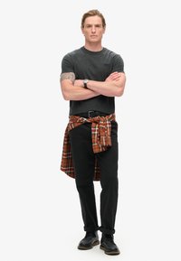 Camiseta gris, pantalones negros y zapatos negros, con una camisa naranja a cuadros atada a la cintura. Figura masculina con los brazos cruzados.