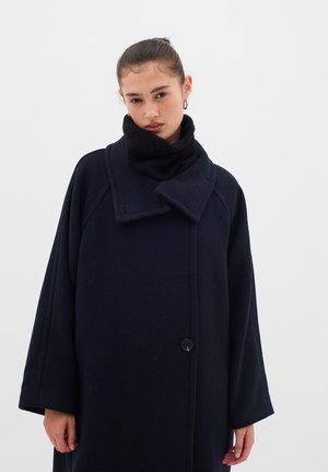 Manteau en laine bleu marine avec un col rabattu, de grandes manches et une fermeture à un bouton. La texture est lisse avec un aspect chaud et confortable.
