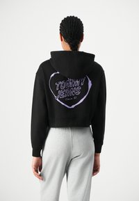 Hoodie nera a taglio corto con una grafica a forma di cuore viola che recita "TOMMY JEANS Since 85" sul retro, abbinata a pantaloni della tuta grigi.